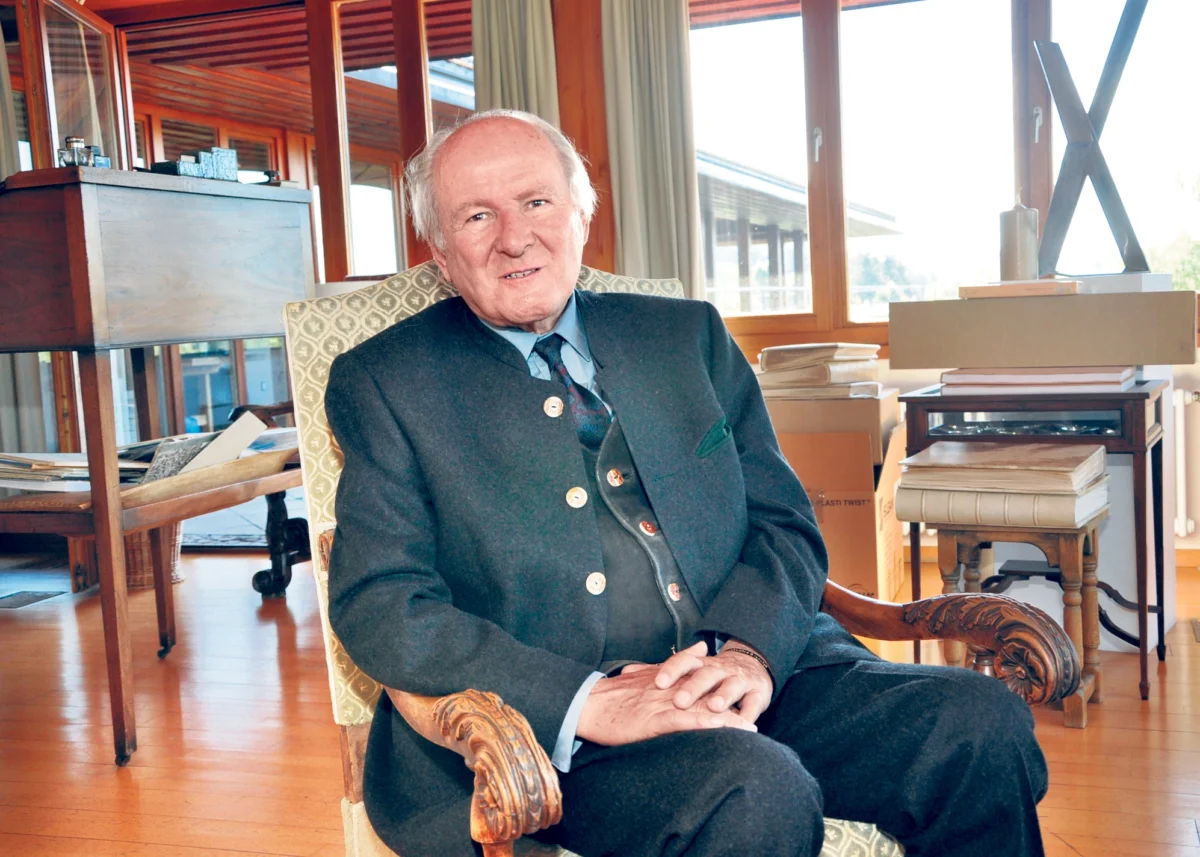 Claus Hipp sitzt in einem Sessel in seinem Arbeitszimmer. Im Hintergrund sind Fensterfronten, Bücherstapel und Holzmöbel zu sehen.