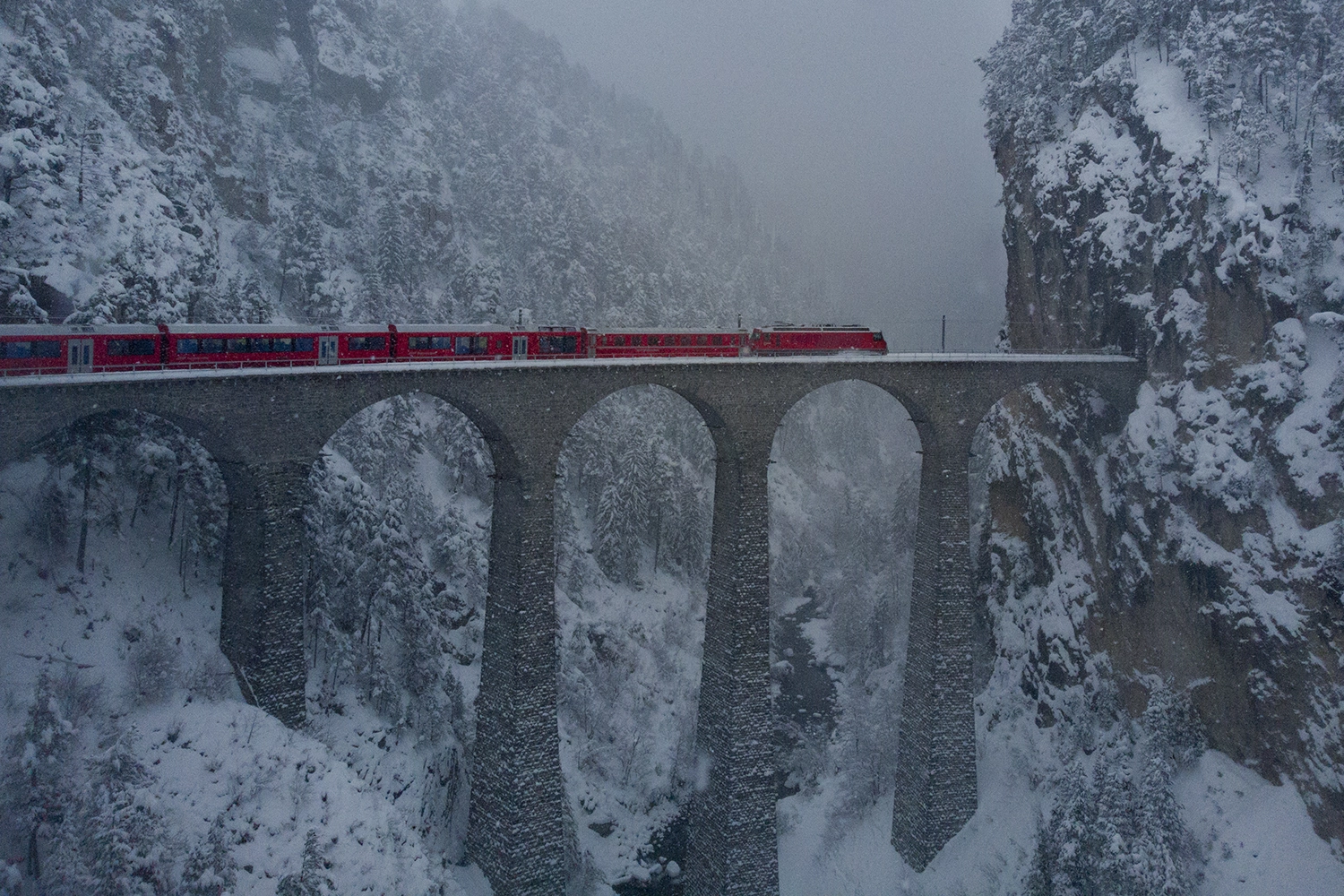 Die Rolle seines Lebens 1 Roter Zug fährt über eine hohe steinerne Brücke mit mehreren Bögen in einer winterlichen, verschneiten Berglandschaft.