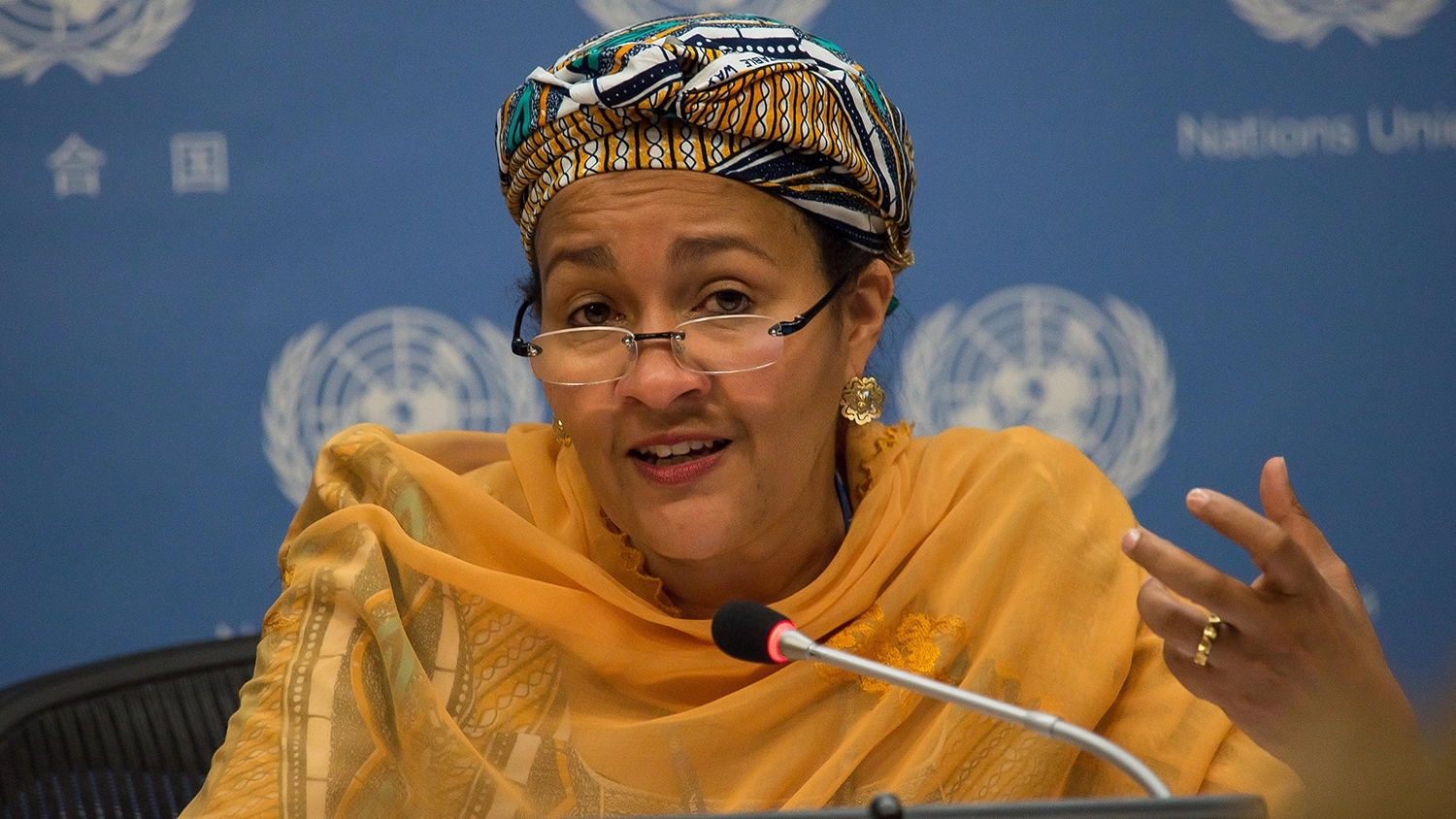 Interview with UN Special Adviser Amina Mohammed 1 Frau in traditioneller afrikanischer Kleidung mit Kopftuch und Brille spricht bei einer Konferenz vor einem blauen Hintergrund mit UN-Logos. Sie gestikuliert mit der Hand und spricht in ein Mikrofon.