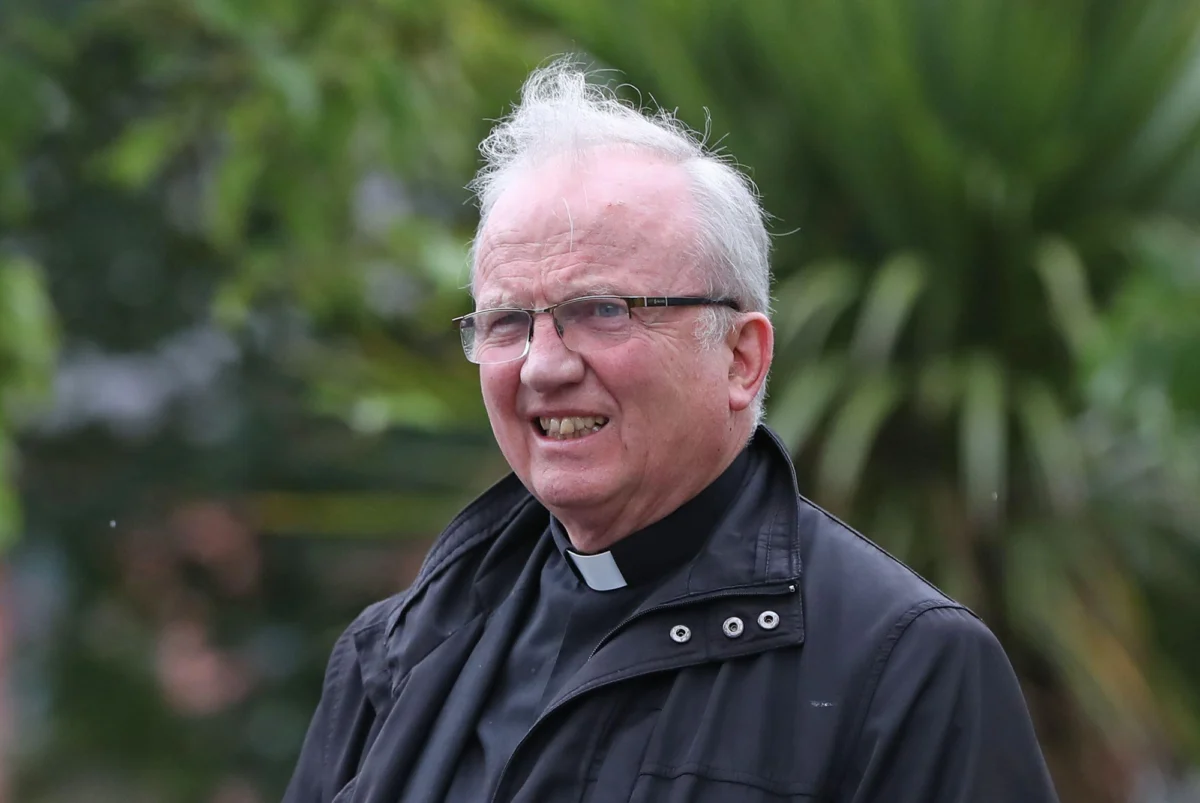 Mann mit Brille und grauen Haaren trägt eine schwarze Jacke und einen Priesterkragen, steht im Freien vor grünem, verschwommenem Hintergrund.