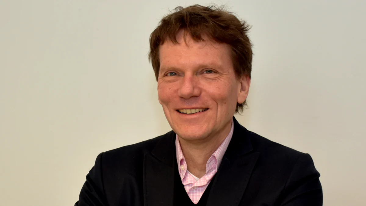Porträt des Soziologen Hartmut Rosa. Er lächelt in die Kamera und trägt ein dunkles Sakko über einem schwarzen Pullover und einem hellrosa Hemd.