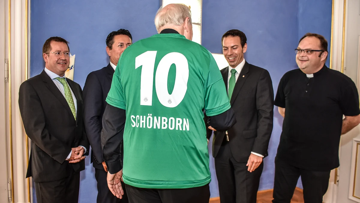 Kardinal Christoph Schönborn trägt ein grünes Rapid-Trikot mit der Rückennummer 10 und seinem Namen, während er mit mehreren Männern in einem festlichen Raum ins Gespräch kommt.