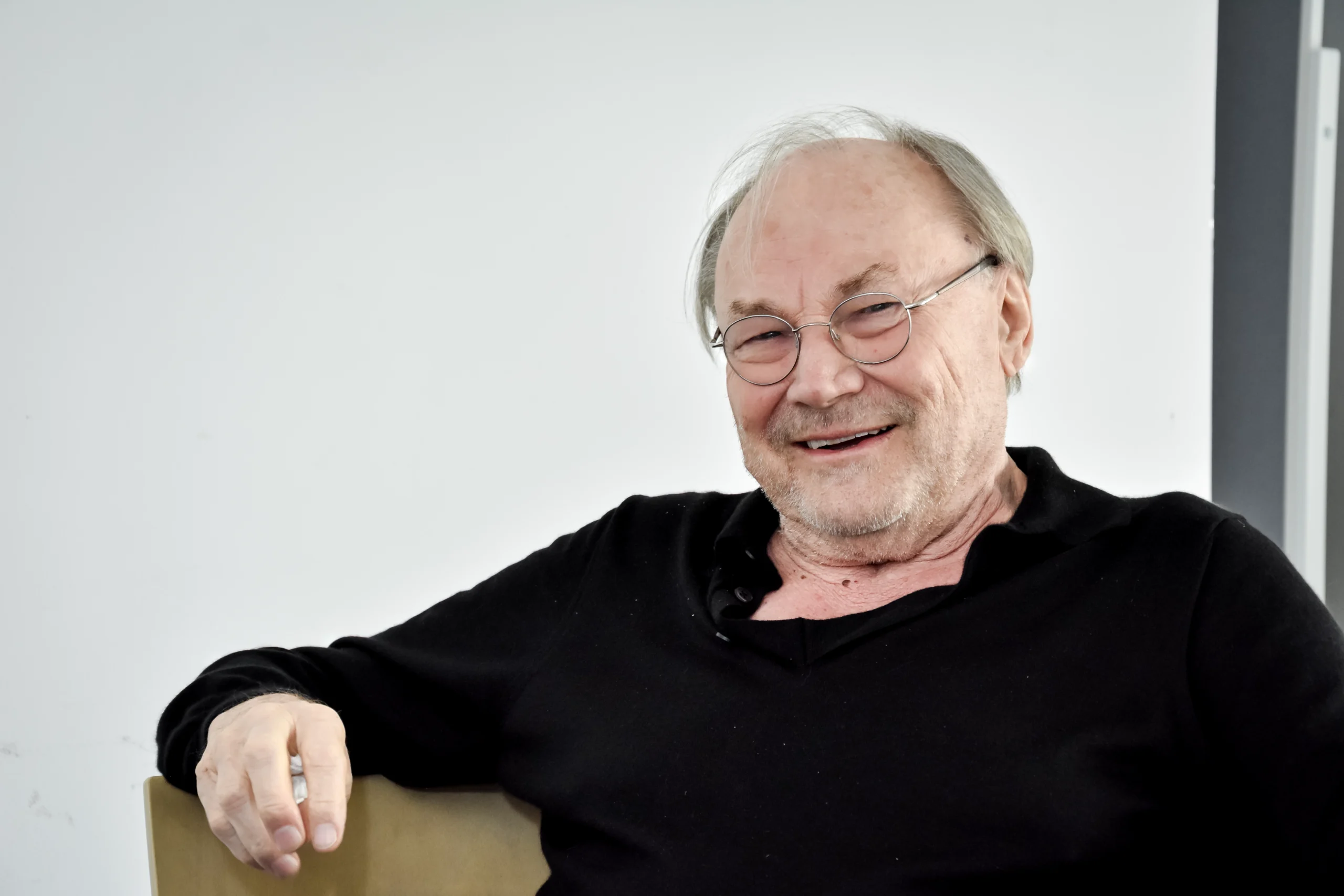 Klaus Maria Brandauer lächelnd im Interview, sitzend mit schwarzem Pullover und Brille.