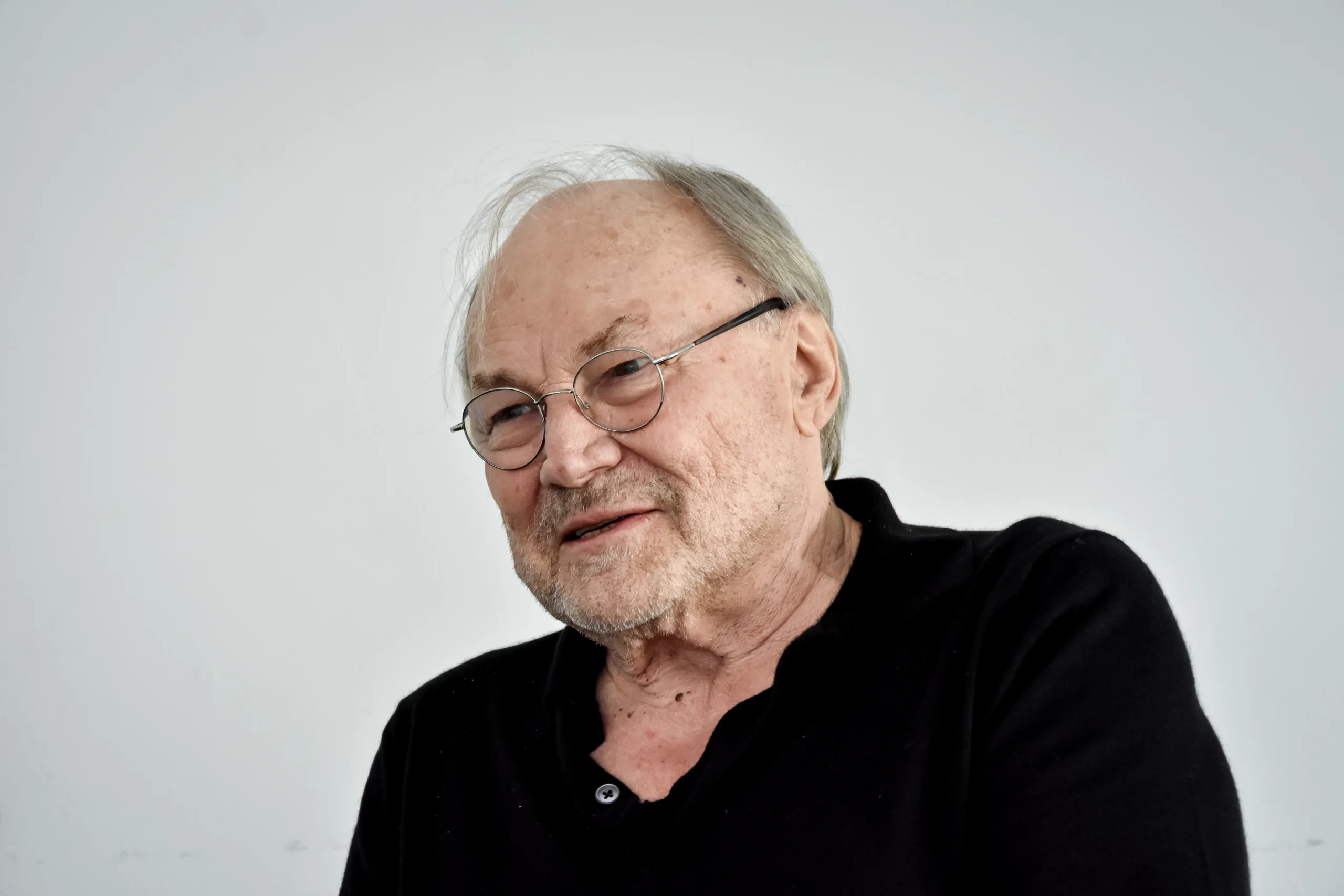 Klaus Maria Brandauer im Gespräch, leicht zur Seite blickend, mit freundlichem Ausdruck, schwarzem Pullover und Brille.