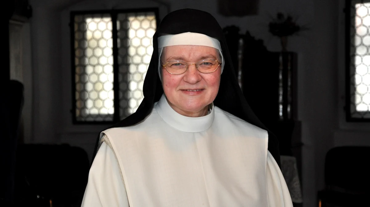 Porträt der Dominikanerin Lydia la Dous in ihrem Ordensgewand. Die ehemalige Astrophysikerin und heutige Theologin sitzt in einem Innenraum vor historischen Fenstern und blickt lächelnd in die Kamera.