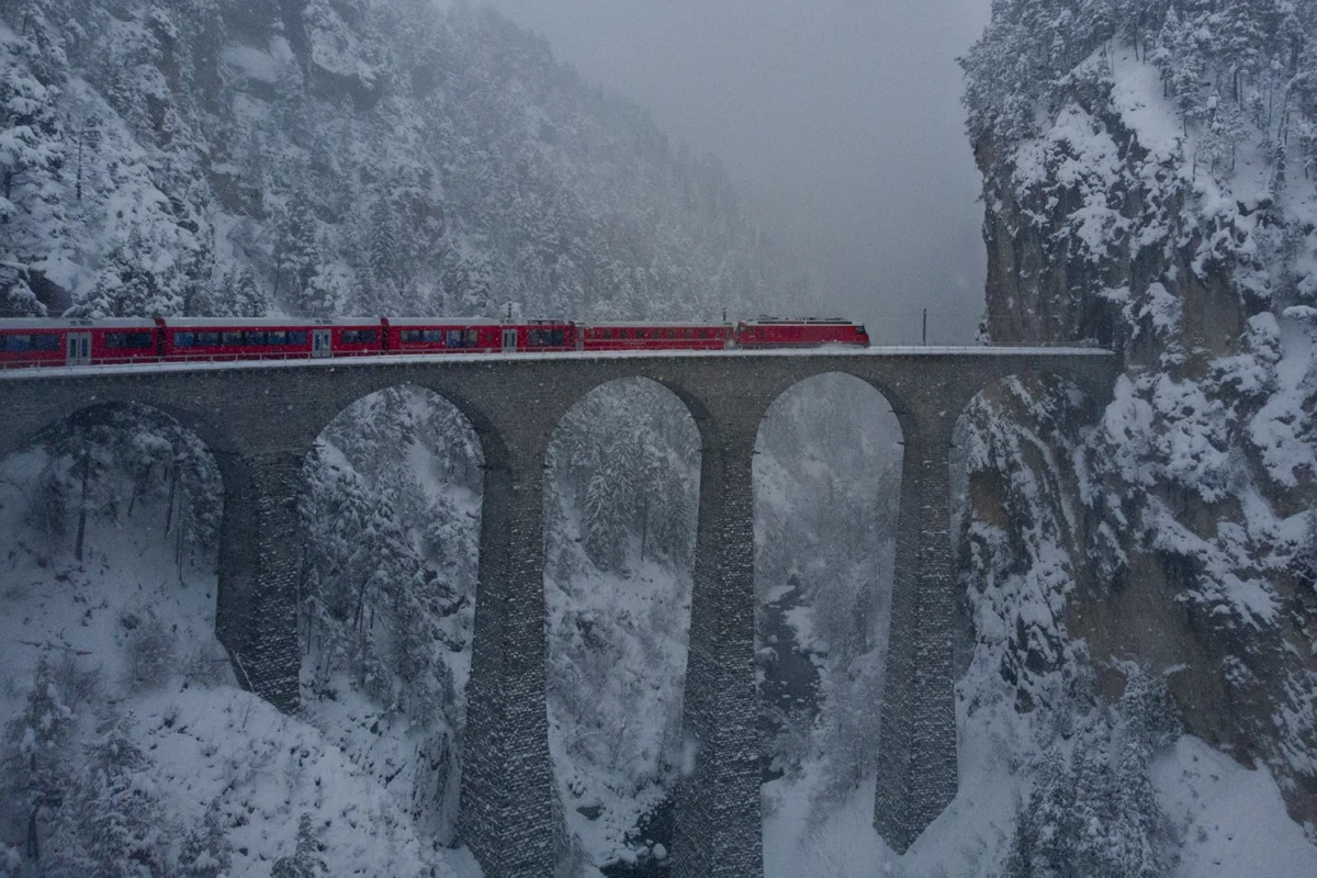 Roter Zug fährt über eine hohe steinerne Brücke mit mehreren Bögen in einer winterlichen, verschneiten Berglandschaft.