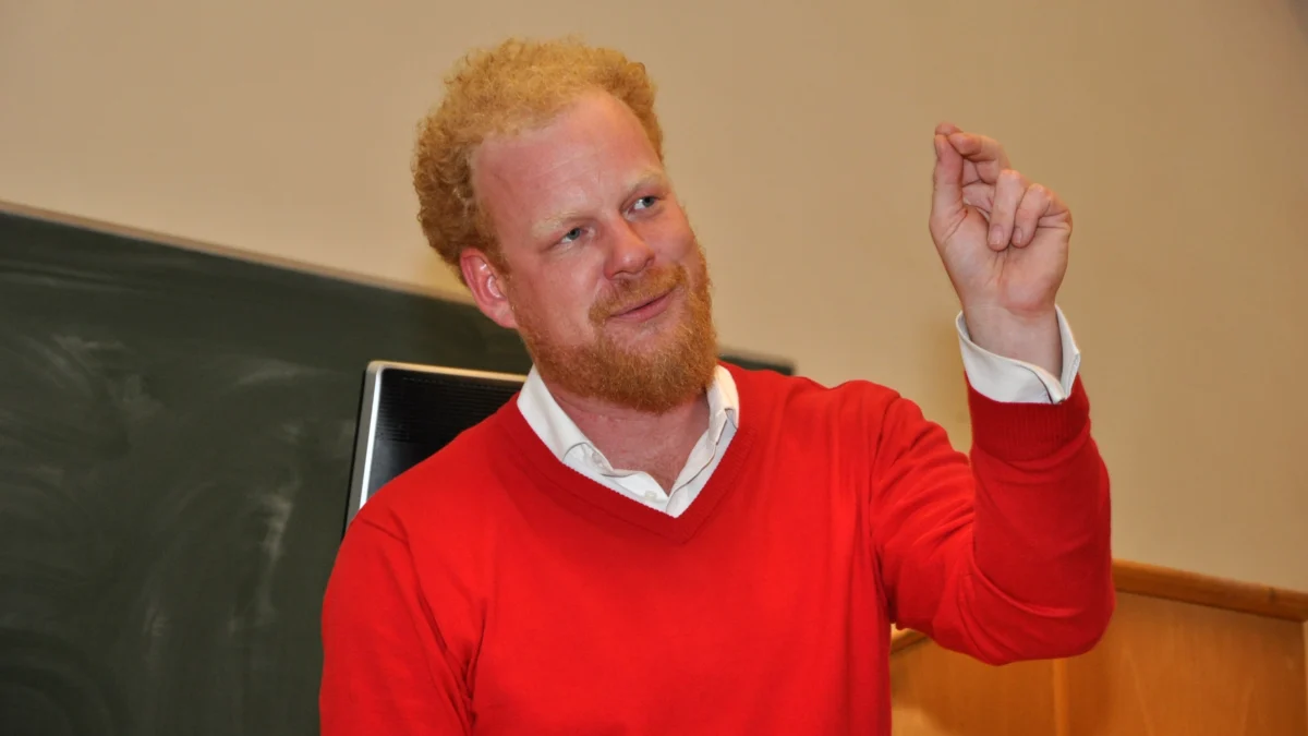 Tomáš Sedláček gesturing during a lecture, wearing a red sweater and white shirt, standing in front of a blackboard.
